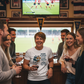 Group of people in a pub watching a rugby match on television. The lady is wearing a Cows Corner t-shirt with the funny rugby slogan 'Rugby Players Are Either Piano Movers or Piano Players – Pierre Danos'. Cows Corner gifts are perfect for sport-mad fans, these gifts work brilliantly for birthdays, new baby celebrations, Father’s Day, Mother’s Day, Christmas, anniversaries, thank you gifts, end-of-season team awards, graduations, retirements, and just-because moments when you want to raise a smile. 
