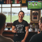 Woman in a rugby-themed shirt laughing in a bar with sports memorabilia on the walls. The lady is wearing a Cows Corner t-shirt with the funny rugby slogan on the front that reads 'Rugby Where Friendly Matches Leave Scars'. Cows Corner gifts are perfect for sport-mad fans, these gifts work brilliantly for birthdays, new baby celebrations, Father’s Day, Mother’s Day, Christmas, anniversaries, thank you gifts, end-of-season team awards, graduations, retirements, and just-because moments when you want to raise