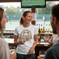People in a bar watching a tennis match on TV, one person wearing a Cows Corner t-shirt with tennis coach slogan on the front that reads 'Behind Every Great Rally Is a Coach Like You – Thank You'. Cows Corner gifts are perfect for sport-mad fans, these gifts work brilliantly for birthdays, new baby celebrations, Father’s Day, Mother’s Day, Christmas, anniversaries, thank you gifts, end-of-season team awards, graduations, retirements, and just-because moments when you want to raise a smile. 
