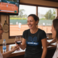 Woman in a black t-shirt with a tennis-themed design, laughing with friends in a room with a tennis court view.  The lady is wearing a Cows Corner T-shirt with a funny tennis slogan on the front that reads 'Game, Set, Mismatch'. Cows Corner gifts are perfect for sport-mad fans, these gifts work brilliantly for birthdays, new baby celebrations, Father’s Day, Mother’s Day, Christmas, anniversaries, thank you gifts, end-of-season team awards, graduations, retirements, and just-because moments.