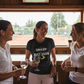 Three women in a sports bar with tennis-themed decor, one wearing a Cows Corner T-shirt with the funny tennis slogan on the front that reads 'I Don't Smash Rackets Just Egos'. Cows Corner gifts are perfect for sport-mad fans, these gifts work brilliantly for birthdays, new baby celebrations, Father’s Day, Mother’s Day, Christmas, anniversaries, thank you gifts, end-of-season team awards, graduations, retirements, and just-because moments when you want to raise a smile. 
