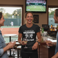 Woman in a sports bar wearing a t-shirt with tennis-themed text, surrounded by friends and a tennis match on TV.  The lady is wearing a Cows Corner T-shirt with the funny tennis slogan on the front that reads 'I've Seen Better Backhands In A Bar Fight'. Cows Corner gifts are perfect for sport-mad fans, these gifts work brilliantly for birthdays, new baby celebrations, Father’s Day, Mother’s Day, Christmas, anniversaries, thank you gifts, end-of-season team awards, graduations, retirements, and just-because.