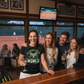 Group of people in a bar with a tennis court visible through the windows. The lady is wearing a Cows Corner t-shirt with the tennis coach slogan on the front that reads 'My Tennis Coach Is the Best in the World (and I’ve Checked)'. Cows Corner gifts are perfect for sport-mad fans, these gifts work brilliantly for birthdays, new baby celebrations, Father’s Day, Mother’s Day, Christmas, anniversaries, thank you gifts, end-of-season team awards, graduations, retirements, and just-because moments 