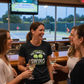 Group of women laughing and holding drinks in a bar with a tennis court visible on the TV. The lady is wearing a Cows Corner t-shirt with the tennis coach slogan on the front that reads 'Thanks Coach You Turned My Swing Into Something Special'. Cows Corner gifts are perfect for sport-mad fans, these gifts work brilliantly for birthdays, new baby celebrations, Father’s Day, Mother’s Day, Christmas, anniversaries, thank you gifts, end-of-season team awards, graduations, retirements, and just-because moments