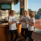 Three women in white t-shirts standing in a room with a tennis court view, holding drinks, all three are wearing a Cows Corner t-shirt with the tennis coach slogan on the front that reads 'Thanks for the Love the Lobs and the Lessons. Best Tennis Coach Ever'. Cows Corner gifts are perfect for sport-mad fans, these gifts work brilliantly for birthdays, new baby celebrations, Father’s Day, Mother’s Day, Christmas, anniversaries, thank you gifts, end-of-season team awards, graduations, retirements