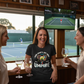 Three women watching a tennis match on a TV screen in a room with a window view of a tennis court.  The lady is wearing a Cows Corner T-shirt with the funny tennis slogan on the front that reads 'Your Game's Got More Faults Than A Bad Umpire' Cows Corner gifts are perfect for sport-mad fans, these gifts work brilliantly for birthdays, new baby celebrations, Father’s Day, Mother’s Day, Christmas, anniversaries, thank you gifts, end-of-season team awards, graduations, retirements, and just-because moments.