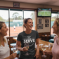 Three women laughing together in a sports bar with a tennis court view.  The lady is wearing a Cows Corner T-shirt with the funny Tennis slogan on the front that reads 'Your Serve Is As Weak As Your Excuses'. Cows Corner gifts are perfect for sport-mad fans, these gifts work brilliantly for birthdays, new baby celebrations, Father’s Day, Mother’s Day, Christmas, anniversaries, thank you gifts, end-of-season team awards, graduations, retirements, and just-because moments when you want to raise a smile.