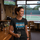 Woman in a bar wearing a t-shirt with humorous text, with tennis court visible in the background.  The lady is wearing a Cows Corner T-shirt with the funny tennis slogan on the front that reads 'Your Serve Is Slower Than My Grandma's Wifi'.  Cows Corner gifts are perfect for sport-mad fans, these gifts work brilliantly for birthdays, new baby celebrations, Father’s Day, Mother’s Day, Christmas, anniversaries, thank you gifts, end-of-season team awards, graduations, retirements, and just-because moments.