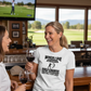 Two women in a golf-themed bar, one wearing a humorous t-shirt. The lady is wearing a Cows Corner t-shirt with a funny golf slogan on the front that reads 'When I Die Bury Me on the Golf Course So My Husband Will Visit'. Cows Corner gifts are perfect for sport-mad fans, these gifts work brilliantly for birthdays, new baby celebrations, Father’s Day, Mother’s Day, Christmas, anniversaries, thank you gifts, end-of-season team awards, graduations, retirements, and just-because moments 