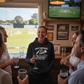 Group of people celebrating a birthday with drinks in a pub, watching cricket on TV. The lady is wearing a Cows Corner hoodie with the cricket birthday slogan that reads 'Happy Birthday! Your balls are officially 'swinging' too low.'. Cows Corner gifts are perfect for sport-mad fans, these gifts work brilliantly for birthdays, new baby celebrations, Father’s Day, Mother’s Day, Christmas, anniversaries, thank you gifts, end-of-season team awards, graduations, retirements, and just-because moments 