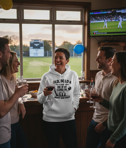 Group of people celebrating a birthday in a sports bar with a TV showing a cricket match. The lady is wearing a Cows Corner hoodie with the funny cricket birthday slogan 'Old, Bold & Still Looking for a Decent Length. Happy Birthday'.  Cows Corner gifts are perfect for sport-mad fans, these gifts work brilliantly for birthdays, new baby celebrations, Father’s Day, Mother’s Day, Christmas, anniversaries, thank you gifts, end-of-season team awards, graduations, retirements, and just-because moments when you w
