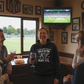 People in a pub watching a sports match on TV, one person wearing a humorous hoodie. The lady is wearing a Cows Corner hoodie with the funny cricket birthday slogan 'You Used to be a Heavy Hitter. Now you’ve Just Got Heavy Tits'. Cows Corner gifts are perfect for sport-mad fans, these gifts work brilliantly for birthdays, new baby celebrations, Father’s Day, Mother’s Day, Christmas, anniversaries, thank you gifts, end-of-season team awards, graduations, retirements, and just-because moments 