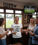 People in a pub watching a sports match on television. The lady is wearing a Cows Corner t-shirt with the funny cricket birthday slogan 'Old, Bold & Still Looking for a Decent Length. Happy Birthday'. Cows Corner gifts are perfect for sport-mad fans, these gifts work brilliantly for birthdays, new baby celebrations, Father’s Day, Mother’s Day, Christmas, anniversaries, thank you gifts, end-of-season team awards, graduations, retirements, and just-because moments when you want to raise a smile. 
