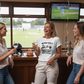 Three women in a sports bar with a TV showing a cricket match, one celebrating a birthday. The lady is wearing a Cows Corner t-shirt with the funny cricket birthday slogan 'Retired Hurt? No, just too old to find the 'Sweet Spot' Happy Birthday'. Cows Corner gifts are perfect for sport-mad fans, these gifts work brilliantly for birthdays, new baby celebrations, Father’s Day, Mother’s Day, Christmas, anniversaries, thank you gifts, end-of-season team awards, graduations, retirements, and just-because moments 