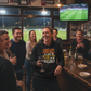 People in a bar watching sports on TV with a person holding a wine glass.  The lady is wearing a Cows Corner t-shirt with the funny fantasy football slogan on the front that reads 'Fantasy Football Is Just Me Getting F*cked Over Weekly'  Cows Corner gifts are perfect for sport-mad fans, these gifts work brilliantly for birthdays, new baby celebrations, Father’s Day, Mother’s Day, Christmas, anniversaries, thank you gifts, end-of-season team awards, graduations, retirements, and just-because moments 