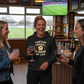 Three women in a pub watching a sports game, one wearing a hoodie with text.  The lady is wearing a Cows Corner hoodie with the funny fantasy football slogan on the front that reads 'Fantasy Football Is Ruining My Life' Cows Corner gifts are perfect for sport-mad fans, these gifts work brilliantly for birthdays, new baby celebrations, Father’s Day, Mother’s Day, Christmas, anniversaries, thank you gifts, end-of-season team awards, graduations, retirements, and just-because moments 