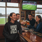 People at a bar with a TV showing a sports game, one person wearing a hoodie with text. The lady is wearing a Cows Corner T-shirt with the funny fantasy football slogan on the front that reads 'I Bench Goals and Start W*nkers'.  Cows Corner gifts are perfect for sport-mad fans, these gifts work brilliantly for birthdays, new baby celebrations, Father’s Day, Mother’s Day, Christmas, anniversaries, thank you gifts, end-of-season team awards, graduations, retirements, and just-because moments 