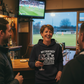 Woman in a bar wearing a hoodie with humorous text, holding a glass of wine, with a sports game on TV in the background.  She is wearing a Cows Corner hoodie with the funny fantasy football slogan on the front that reads 'My Fantasy Team Is F*cking Useless'.  Cows Corner gifts are perfect for sport-mad fans, these gifts work brilliantly for birthdays, new baby celebrations, Father’s Day, Mother’s Day, Christmas, anniversaries, thank you gifts, end-of-season team awards, graduations, retirements