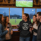 Group of people in a sports bar watching a soccer game on TV.  The lady is wearing a Cows Corner hoodie with the funny fantasy football slogan on the front that reads 'Talk Big, Play Sh*t, Finish Last'. Cows Corner gifts are perfect for sport-mad fans, these gifts work brilliantly for birthdays, new baby celebrations, Father’s Day, Mother’s Day, Christmas, anniversaries, thank you gifts, end-of-season team awards, graduations, retirements, and just-because moments when you want to raise a smile. 