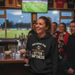 Woman in a bar wearing a humorous hoodie with a soccer game on TV in the background.  The lady is wearing a Cows Corner hoodie with the funny fantasy football slogan on the front that reads 'Your Fantasy Team Has One Setting: Utter Sh*tness'. Cows Corner gifts are perfect for sport-mad fans, these gifts work brilliantly for birthdays, new baby celebrations, Father’s Day, Mother’s Day, Christmas, anniversaries, thank you gifts, end-of-season team awards, graduations, retirements, and just-because moments