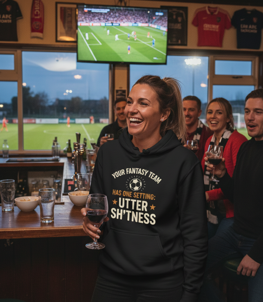 Woman in a bar wearing a humorous hoodie with a soccer game on TV in the background.  The lady is wearing a Cows Corner hoodie with the funny fantasy football slogan on the front that reads 'Your Fantasy Team Has One Setting: Utter Sh*tness'. Cows Corner gifts are perfect for sport-mad fans, these gifts work brilliantly for birthdays, new baby celebrations, Father’s Day, Mother’s Day, Christmas, anniversaries, thank you gifts, end-of-season team awards, graduations, retirements, and just-because moments