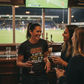 Three women in a sports bar watching a football game on TV.  The lady is wearing a Cows Corner t-shirt with the funny fantasy football slogan on the front that reads 'Fantasy Football Is Just Me Getting F*cked Over Weekly'. Cows Corner gifts are perfect for sport-mad fans, these gifts work brilliantly for birthdays, new baby celebrations, Father’s Day, Mother’s Day, Christmas, anniversaries, thank you gifts, end-of-season team awards, graduations, retirements, and just-because moments 