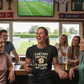 Group of people in a pub watching a soccer game on TV, with one person wearing a 'Fantasy Football is Ruining My Life' shirt.  Cows Corner gifts are perfect for sport-mad fans, these gifts work brilliantly for birthdays, new baby celebrations, Father’s Day, Mother’s Day, Christmas, anniversaries, thank you gifts, end-of-season team awards, graduations, retirements, and just-because moments when you want to raise a smile. Designed for people who appreciate thoughtful gifting with a playful edge