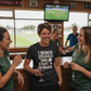 Group of women in a sports bar watching a game on TV, one wearing a humorous t-shirt.  The lady is wearing a Cows Corner T-shirt with the funny fantasy football slogan on the front that reads 'I Bench Goals and Start W*nkers'. Cows Corner gifts are perfect for sport-mad fans, these gifts work brilliantly for birthdays, new baby celebrations, Father’s Day, Mother’s Day, Christmas, anniversaries, thank you gifts, end-of-season team awards, graduations, retirements, and just-because moments 