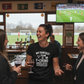 People in a pub watching sports on television with one person wearing a humorous t-shirt.  The lady is wearing a Cows Corner hoodie with the funny fantasy football slogan on the front that reads 'My Fantasy Team Is F*cking Useless'.  Cows Corner gifts are perfect for sport-mad fans, these gifts work brilliantly for birthdays, new baby celebrations, Father’s Day, Mother’s Day, Christmas, anniversaries, thank you gifts, end-of-season team awards, graduations, retirements, and just-because moments 