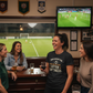 Group of women watching a soccer game on TV in a bar.  The lady is wearing a Cows Corner t-shirt with the funny fantasy football slogan on the front that reads 'Your Fantasy Team Has One Setting: Utter Sh*tness'. Cows Corner gifts are perfect for sport-mad fans, these gifts work brilliantly for birthdays, new baby celebrations, Father’s Day, Mother’s Day, Christmas, anniversaries, thank you gifts, end-of-season team awards, graduations, retirements, and just-because moments when you want to raise a smile. 