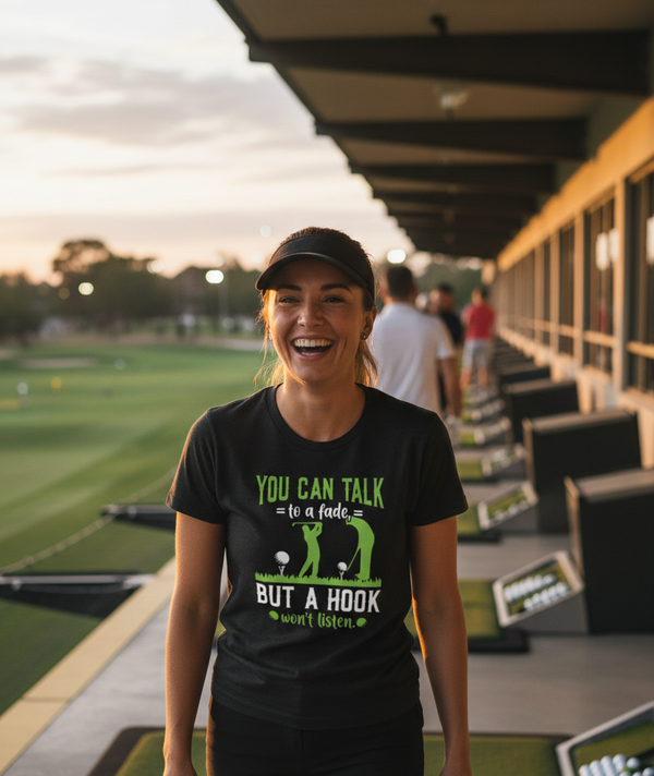 Woman wearing a black t-shirt with a humorous golf graphic and text, standing on a golf course. The lady is wearing a Cows Corner t-shirt with the funny golf slogan 'You Can Talk to a Fade but a Hook Won’t Listen'. Cows Corner gifts are perfect for sport-mad fans, these gifts work brilliantly for birthdays, new baby celebrations, Father’s Day, Mother’s Day, Christmas, anniversaries, thank you gifts, end-of-season team awards, graduations, retirements, and just-because moments when you want to raise a smile.