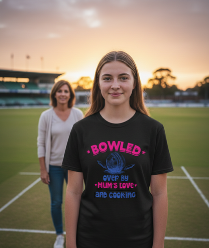 Girl wearing a black t-shirt with colorful text on a sports field at sunset. The girl is wearing a Cows Corner t-shirt with the funny cricket slogan 'Bowled Over by My Mum’s Love & Cooking'. Cows Corner gifts are perfect for sport-mad fans, these gifts work brilliantly for birthdays, new baby celebrations, Father’s Day, Mother’s Day, Christmas, anniversaries, thank you gifts, end-of-season team awards, graduations, retirements, and just-because moments when you want to raise a smile. 
