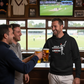 Three men in a pub watching a cricket match on television, clinking beer glasses. The man is wearing a Cows Corner hoodie with the funny cricket slogan 'Bowled & Bamboozled and still looking better than you'.  Cows Corner gifts are perfect for sport-mad fans, these gifts work brilliantly for birthdays, new baby celebrations, Father’s Day, Mother’s Day, Christmas, anniversaries, thank you gifts, end-of-season team awards, graduations, retirements, and just-because moments when you want to raise a smile. 
