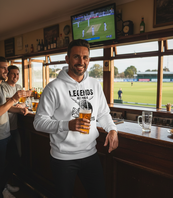 Man in a white hoodie holding a beer in a pub with a cricket match on TV. The man is wearing a Cows Corner hoodie with the funny cricket slogan that reads 'Legends Are Made in the Nets'. Cows Corner gifts are perfect for sport-mad fans, these gifts work brilliantly for birthdays, new baby celebrations, Father’s Day, Mother’s Day, Christmas, anniversaries, thank you gifts, end-of-season team awards, graduations, retirements, and just-because moments when you want to raise a smile. 
