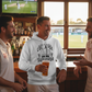 Three men in sports attire, one holding a beer, in a bar setting with a cricket match on TV. The man is wearing a Cows Corner hoodie with a funny cricket slogan 'Middle-Order Collapse Survivor'. Cows Corner gifts are perfect for sport-mad fans, these gifts work brilliantly for birthdays, new baby celebrations, Father’s Day, Mother’s Day, Christmas, anniversaries, thank you gifts, end-of-season team awards, graduations, retirements, and just-because moments when you want to raise a smile. 
