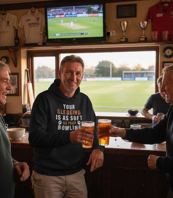 Three men in a pub with a television showing a sports match, holding drinks and smiling. The man is wearing a Cows Corner hoodie with the funny cricket slogan that reads 'Your Sledging Is As Soft As Your Bowling'. Cows Corner gifts are perfect for sport-mad fans, these gifts work brilliantly for birthdays, new baby celebrations, Father’s Day, Mother’s Day, Christmas, anniversaries, thank you gifts, end-of-season team awards, graduations, retirements, and just-because moments when you want to raise a smile. 