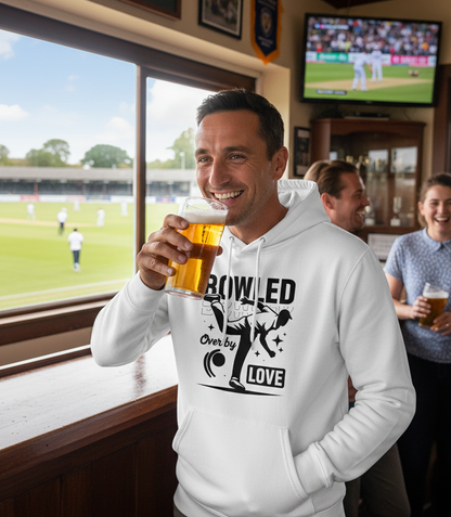 Man in a white hoodie with a humorous design, holding a beer in a pub setting. The man is wearing a Cows Corner hoodie with the funny cricket slogan 'Bowled Over by Love'. Cows Corner gifts are perfect for sport-mad fans, these gifts work brilliantly for birthdays, new baby celebrations, Father’s Day, Mother’s Day, Christmas, anniversaries, thank you gifts, end-of-season team awards, graduations, retirements, and just-because moments when you want to raise a smile. 
