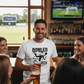 Man in a bar wearing a t-shirt with a sports-themed design, surrounded by friends.  The man is wearing a Cows Corner T-shirt with the funny cricket slogan on the front that reads 'Bowled Over by Love'.  Cows Corner gifts are perfect for sport-mad fans, these gifts work brilliantly for birthdays, new baby celebrations, Father’s Day, Mother’s Day, Christmas, anniversaries, thank you gifts, end-of-season team awards, graduations, retirements, and just-because moments when you want to raise a smile. 