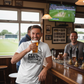 Man in a bar holding a beer, wearing a Cows Corner T-shirt with a funny cricket slogan on the front that reads 'Club Cricket where Everyone’s an All-Rounder'.  Cows Corner gifts are perfect for sport-mad fans, these gifts work brilliantly for birthdays, new baby celebrations, Father’s Day, Mother’s Day, Christmas, anniversaries, thank you gifts, end-of-season team awards, graduations, retirements, and just-because moments when you want to raise a smile. 