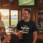 Man in a bar wearing a black t-shirt with a humorous quote, holding a beer, with another person partially visible, the man is wearing a Cows Corner funny t-shirt with the slogan on the front 'I play better with a stiff handle'