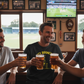 Three men in a pub are clinking beer glasses with a cricket game on a screen in the background.  The man is wearing a Cows Corner t-shirt with the funny cricket slogan on the front saying 'Nice Bouncers'