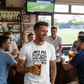 Man wearing a t-shirt with a humorous message, surrounded by friends in a pub watching sports on TV.  The man is wearing a Cows Corner T-shirt with the funny cricket slogan on the front that reads 'Not All Heroes Wear Capes, Some Wear Pads.' Cows Corner gifts are perfect for sport-mad fans, these gifts work brilliantly for birthdays, new baby celebrations, Father’s Day, Mother’s Day, Christmas, anniversaries, thank you gifts, end-of-season team awards, graduations, retirements, and just-because moments.