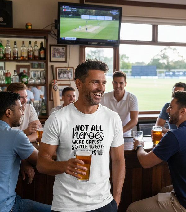 Man wearing a t-shirt with a humorous message, surrounded by friends in a pub watching sports on TV.  The man is wearing a Cows Corner T-shirt with the funny cricket slogan on the front that reads 'Not All Heroes Wear Capes, Some Wear Pads.' Cows Corner gifts are perfect for sport-mad fans, these gifts work brilliantly for birthdays, new baby celebrations, Father’s Day, Mother’s Day, Christmas, anniversaries, thank you gifts, end-of-season team awards, graduations, retirements, and just-because moments.