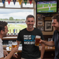 Three men in a pub watching a sports match on TV, one wearing a humorous t-shirt.  The man is wearing a Cows Corner t-shirt with the funny cricket slogan on the front saying 'Pad Up, Shut Up And Try Not To Nick Off'