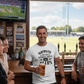 Man holding a beer in a pub with a TV showing cricket and people around him.  The man is wearing a Cows Corner T-shirt with the funny cricket slogan on the front that reads 'Sundays Are for Sledges, Sixes & Beers'.  Cows Corner gifts are perfect for sport-mad fans, these gifts work brilliantly for birthdays, new baby celebrations, Father’s Day, Mother’s Day, Christmas, anniversaries, thank you gifts, end-of-season team awards, graduations, retirements, and just-because moments when you want to raise a smile