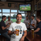Man in a pub holding a beer with a TV showing a sports game in the background. The man is wearing a Cows Corner t-shirt with the cricket coach slogan on the front that reads ;To My Coach Who Made Me Believe in My First Cover Drive – You’re a Legend. Cows Corner gifts are perfect for sport-mad fans, these gifts work brilliantly for birthdays, new baby celebrations, Father’s Day, Mother’s Day, Christmas, anniversaries, thank you gifts, end-of-season team awards, graduations, retirements, and just-because