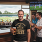 Man in a bar wearing a black t-shirt with a humorous cricket-themed design, holding a beer.  The man is wearing a Cows Corner t-shirt with the funny cricket saying on the front saying 'You Bring The Sledging, I'll Bring The Sixes'