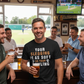 Man in a pub wearing a humorous t-shirt with a group of people around him.  The man is wearing a Cows Corner T-shirt with the funny cricket slogan on the front saying 'Your Sledging Is As Soft As Your Bowling