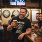 Man in a bar holding a beer, wearing a t-shirt with a dartboard design, surrounded by people and dartboards on the wall. The man is wearing a Cows Corner t-shirt with the funny darts slogan 'Darts Because Therapy is Expensive'. Cows Corner gifts are perfect for sport-mad fans, these gifts work brilliantly for birthdays, new baby celebrations, Father’s Day, Mother’s Day, Christmas, anniversaries, thank you gifts, end-of-season team awards, graduations, retirements, and just-because moments 