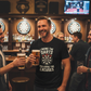 Man in a bar holding a beer and a dartboard-themed t-shirt with a humorous message. The man is wearing a Cows Corner t-shirt with the funny darts slogan 'I Bring the Darts You Bring the Excuses'. Cows Corner gifts are perfect for sport-mad fans, these gifts work brilliantly for birthdays, new baby celebrations, Father’s Day, Mother’s Day, Christmas, anniversaries, thank you gifts, end-of-season team awards, graduations, retirements, and just-because moments when you want to raise a smile. 
